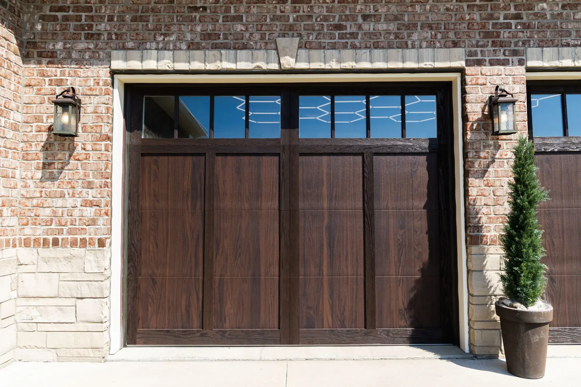 Dark woodtone stamped carriage-house garage door with a row of top windows on a brick home with a stone base