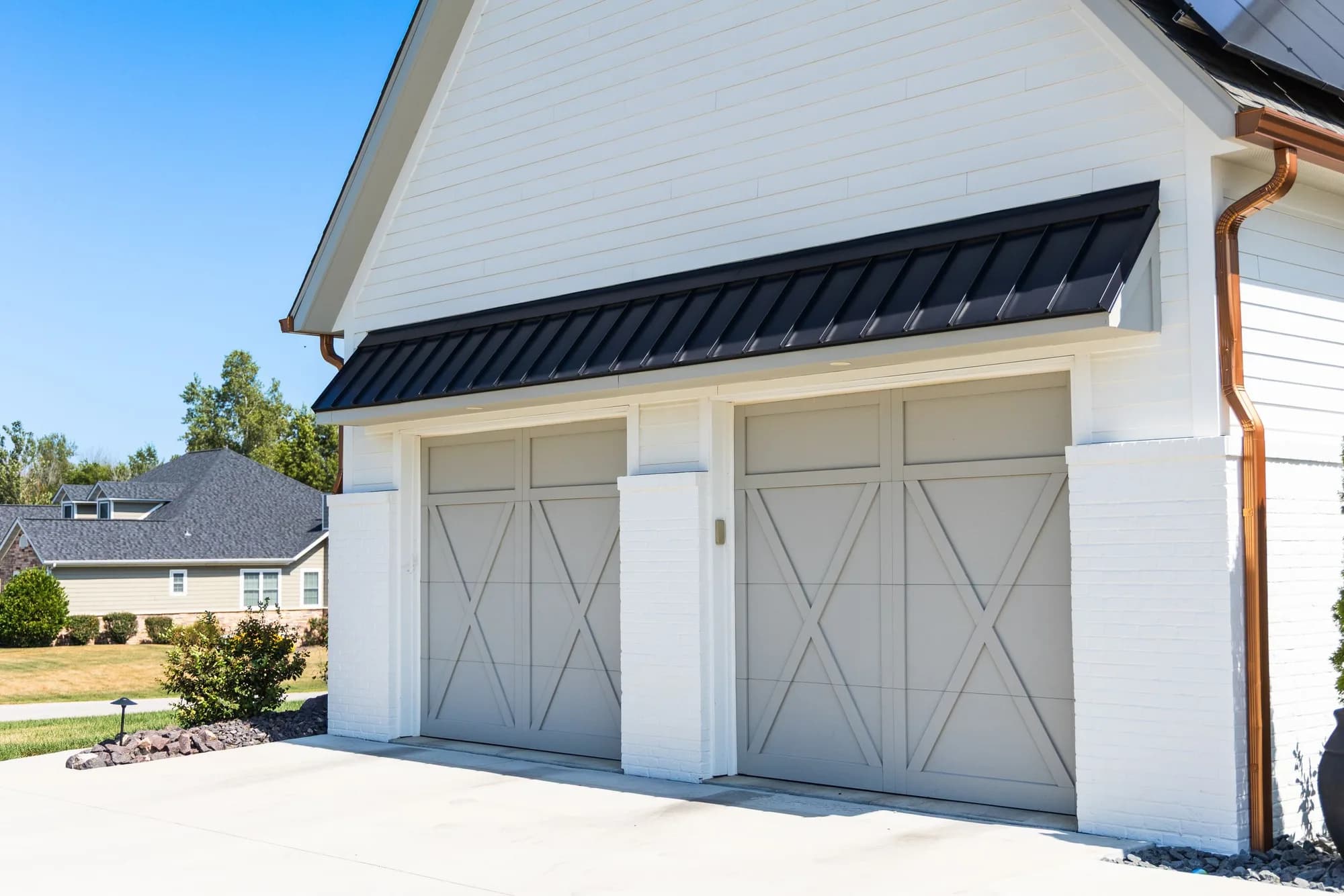 Two light grey carriage overlay garage doors with X-brace boards under a black metal awning on a white painted brick garage