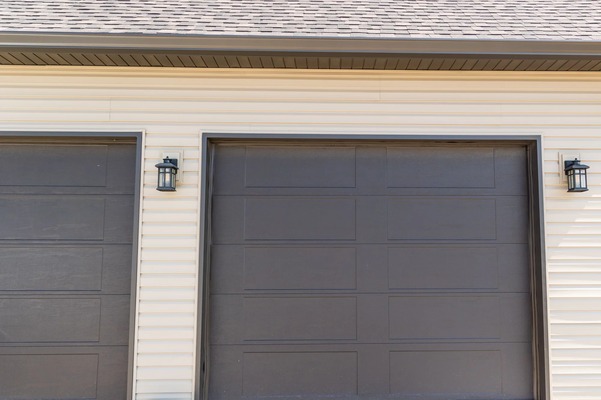 Dark bronze recessed long-panel garage doors on a tan vinyl-sided home with coach lanterns