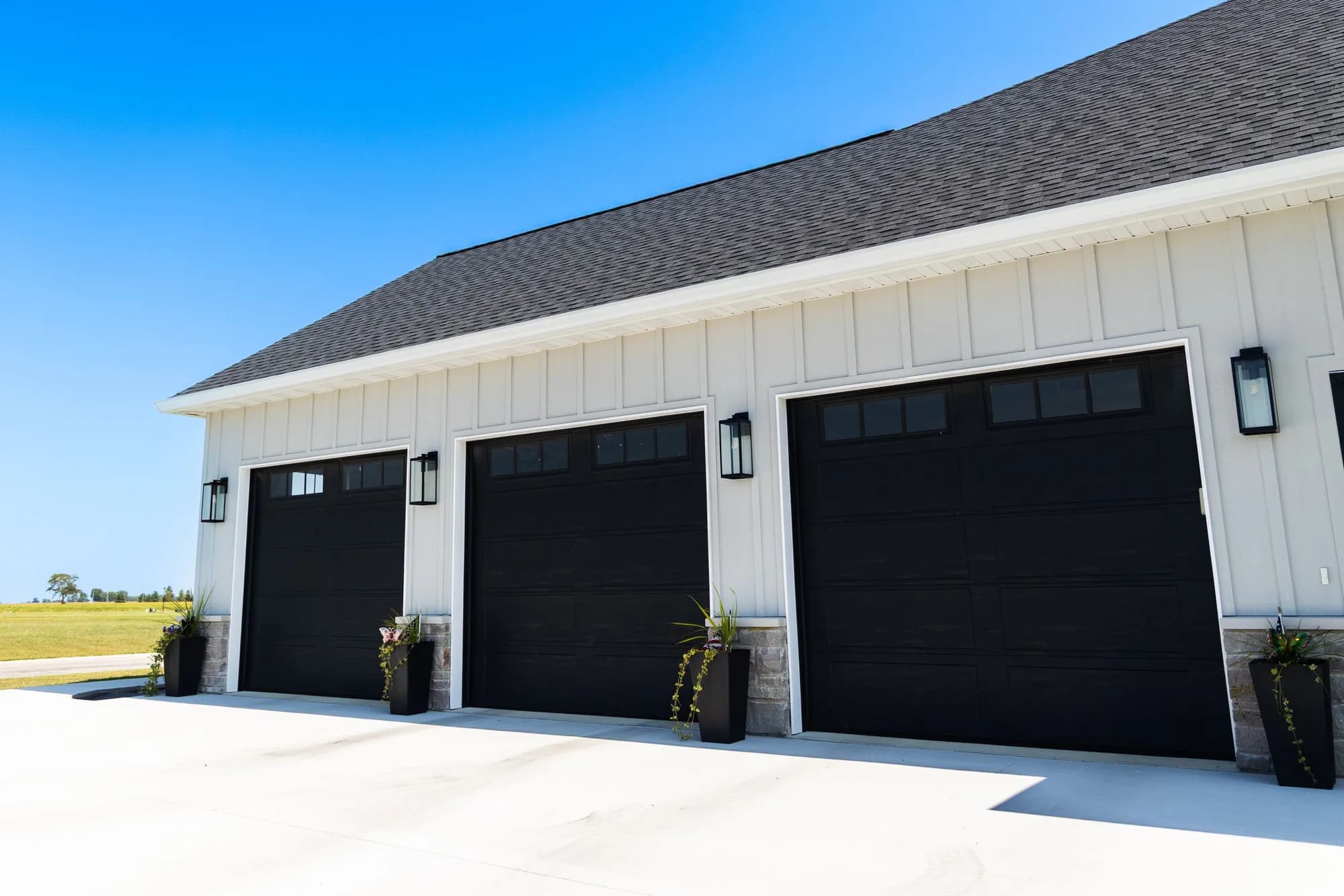 Three black garage doors with top windows on a white farmhouse garage, seen from the driveway